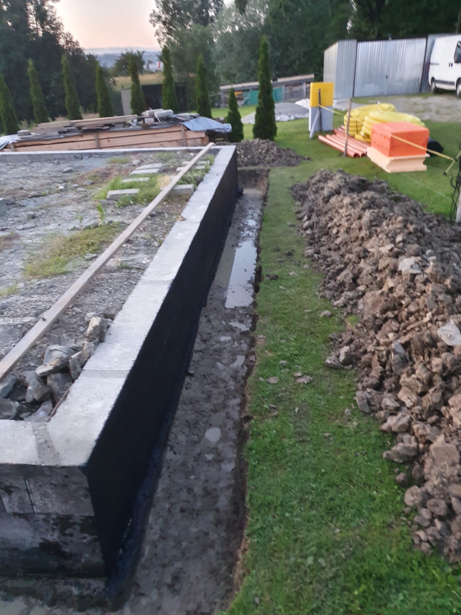Betonowa podmurówka fundamentowa z hydroizolacją pionową w wykopie, widoczne materiały budowlane, ziemia i trawa na działce.