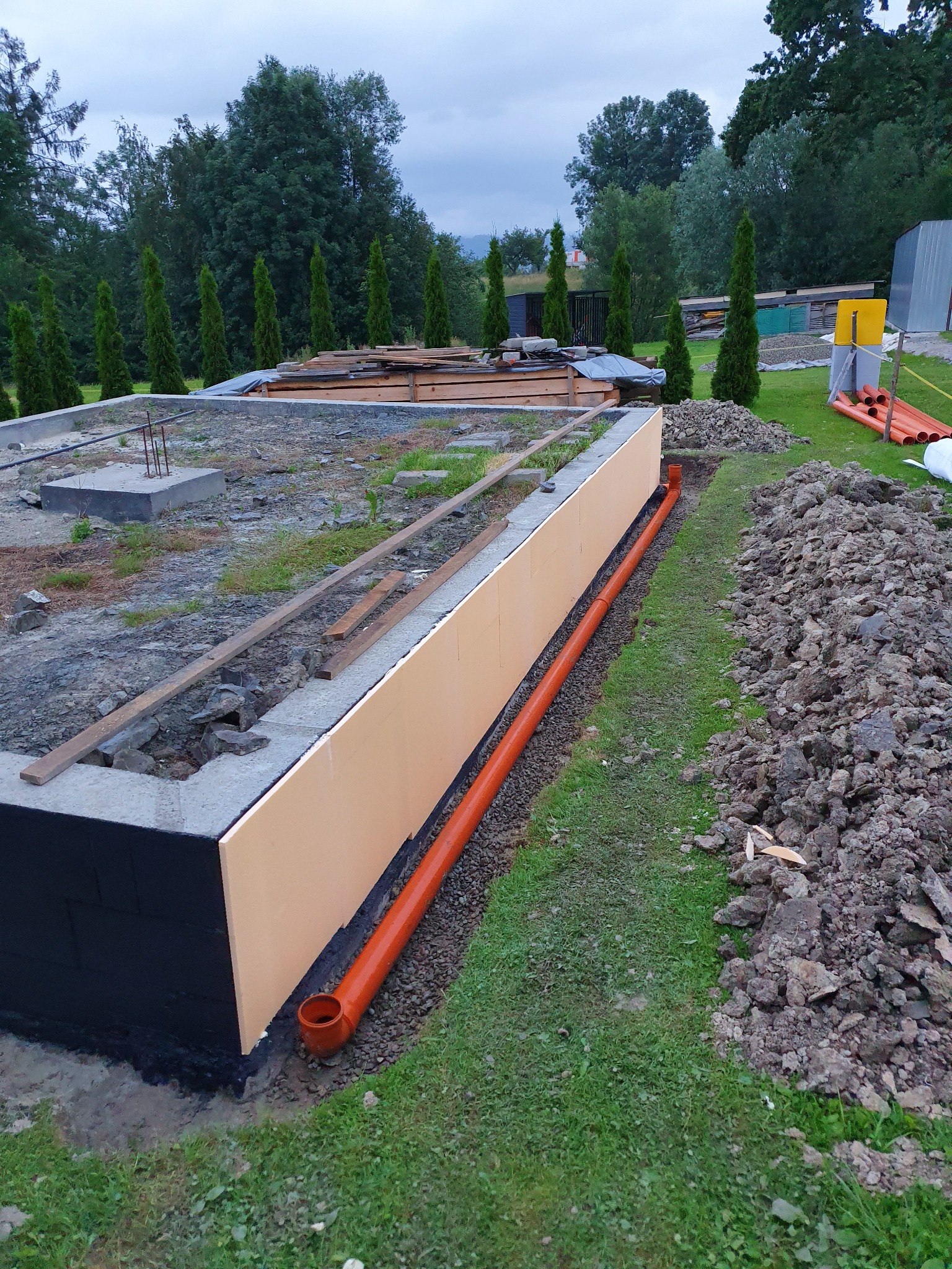 Fundament budynku z izolacją termiczną w kolorze beżowym, zabezpieczony czarną hydroizolacją, z ułożoną rurą drenarską w wykopie, na tle trawy i pryzmy ziemi.