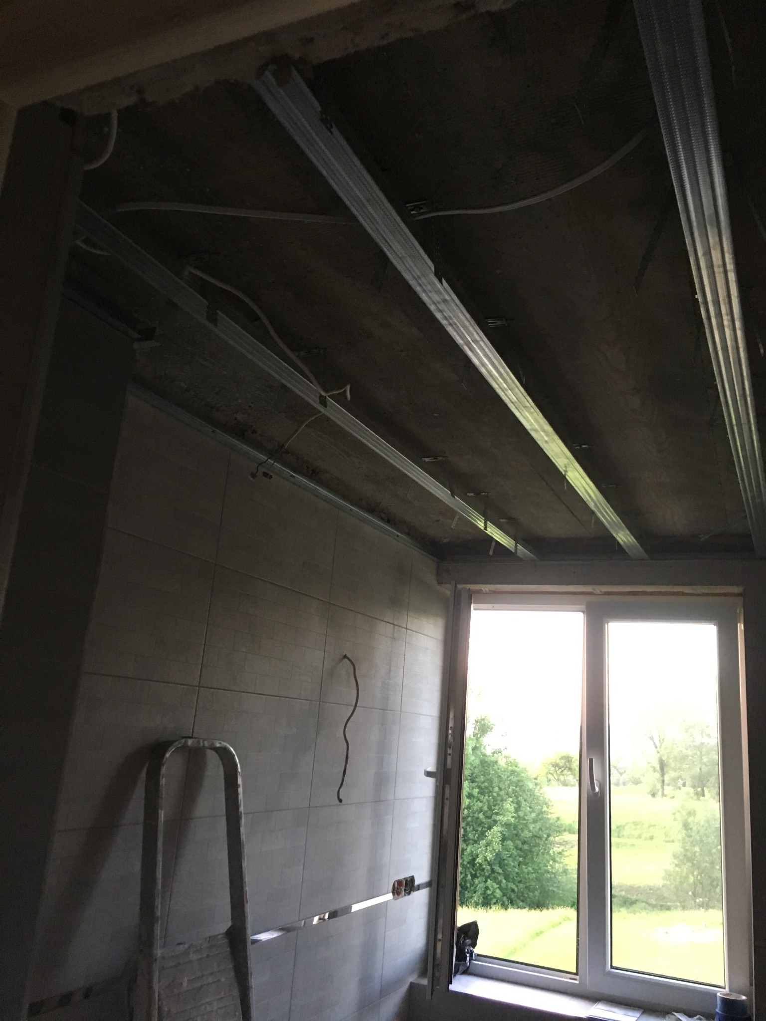 Metalowy stelaż podwieszanego sufitu zamontowany na tle surowego stropu, widoczne przewody elektryczne, płytki ścienne, drabina i okno z widokiem na zieleń.