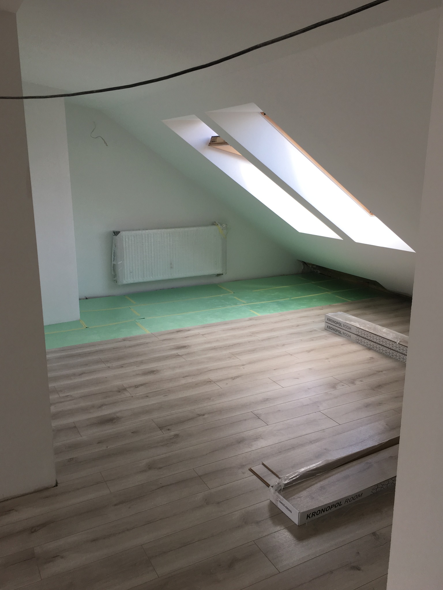 Poddasze w trakcie układania paneli podłogowych Kronopol Room, widoczny fragment ocieplonej podłogi, okno dachowe i zakryty folią grzejnik.