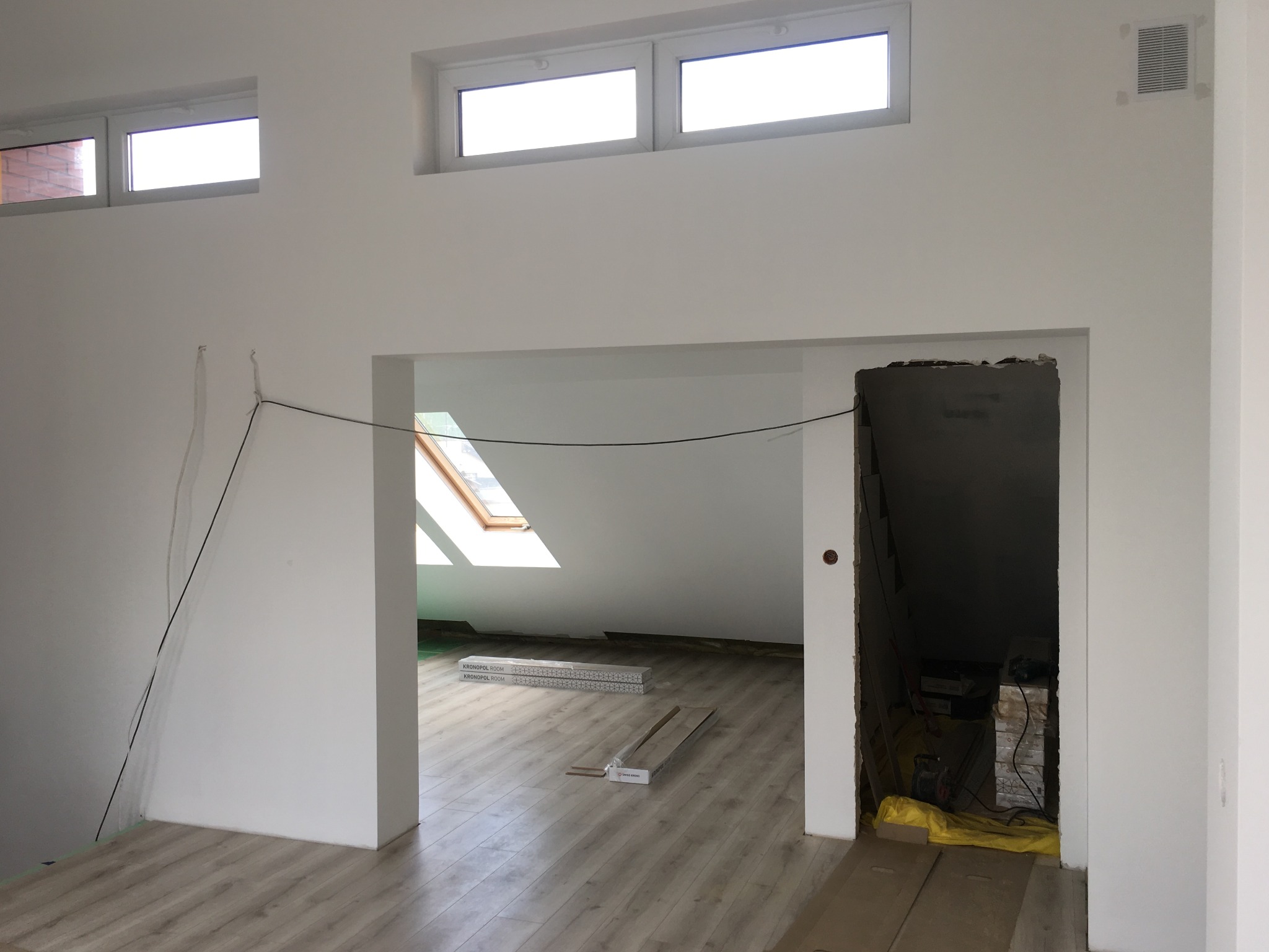 Poddasze w trakcie prac wykończeniowych: widoczne białe ściany, jasne panele podłogowe, okno dachowe, przygotowane paczki paneli oraz otwór w ścianie z widocznym pomieszczeniem gospodarczym...