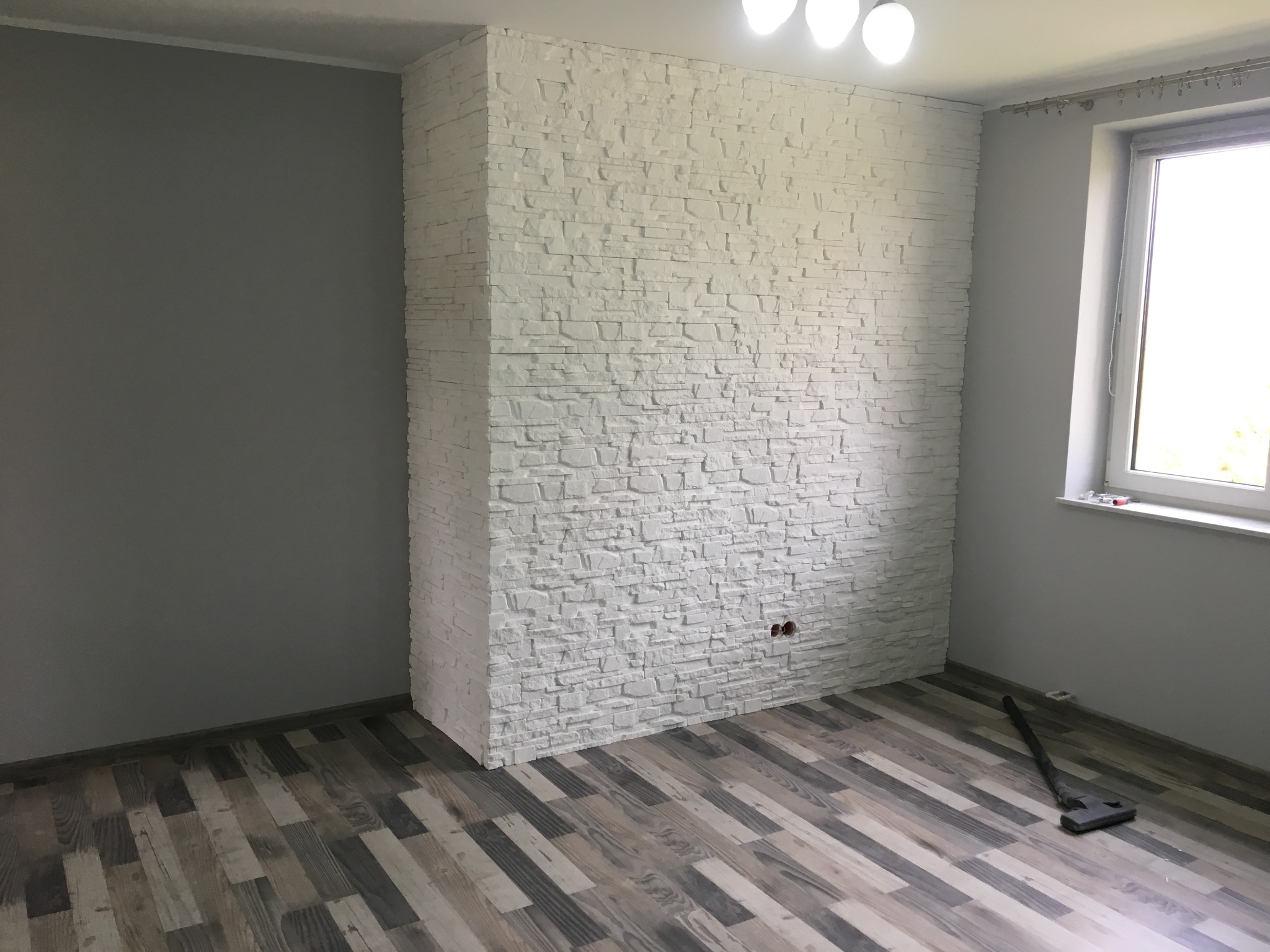 Ściana wykończona białą cegłą dekoracyjną, z widocznymi gniazdkami elektrycznymi, na podłodze panele w odcieniach szarości i brązu, odkurzacz w rogu pokoju, szara ściana boczna i okno z roletą.