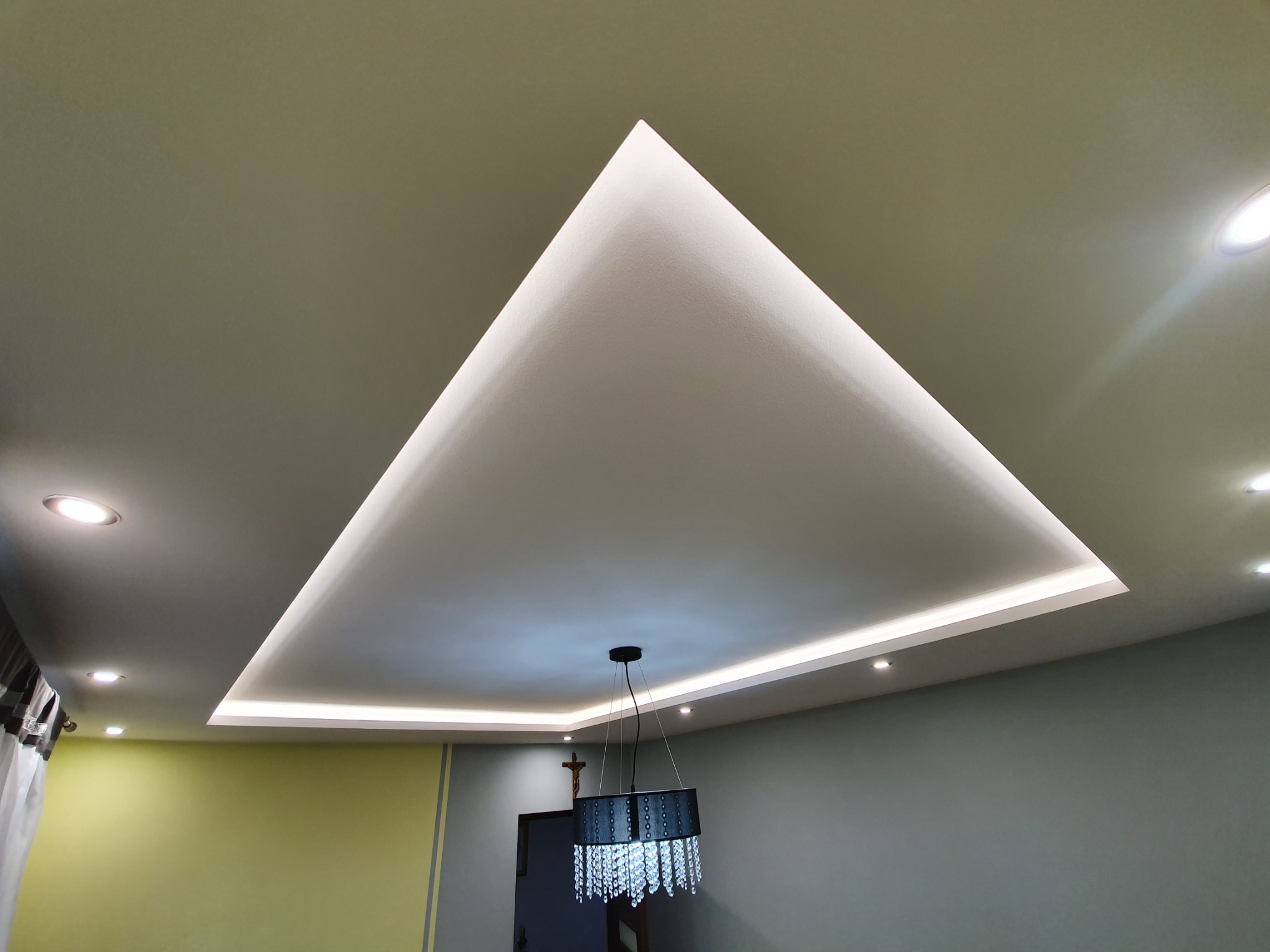 Nowoczesny sufit z trójkątną wnęką podświetloną taśmą LED, w tle widoczny fragment ściany w dwóch odcieniach szarości i żółci oraz wisząca lampa z kryształkami.