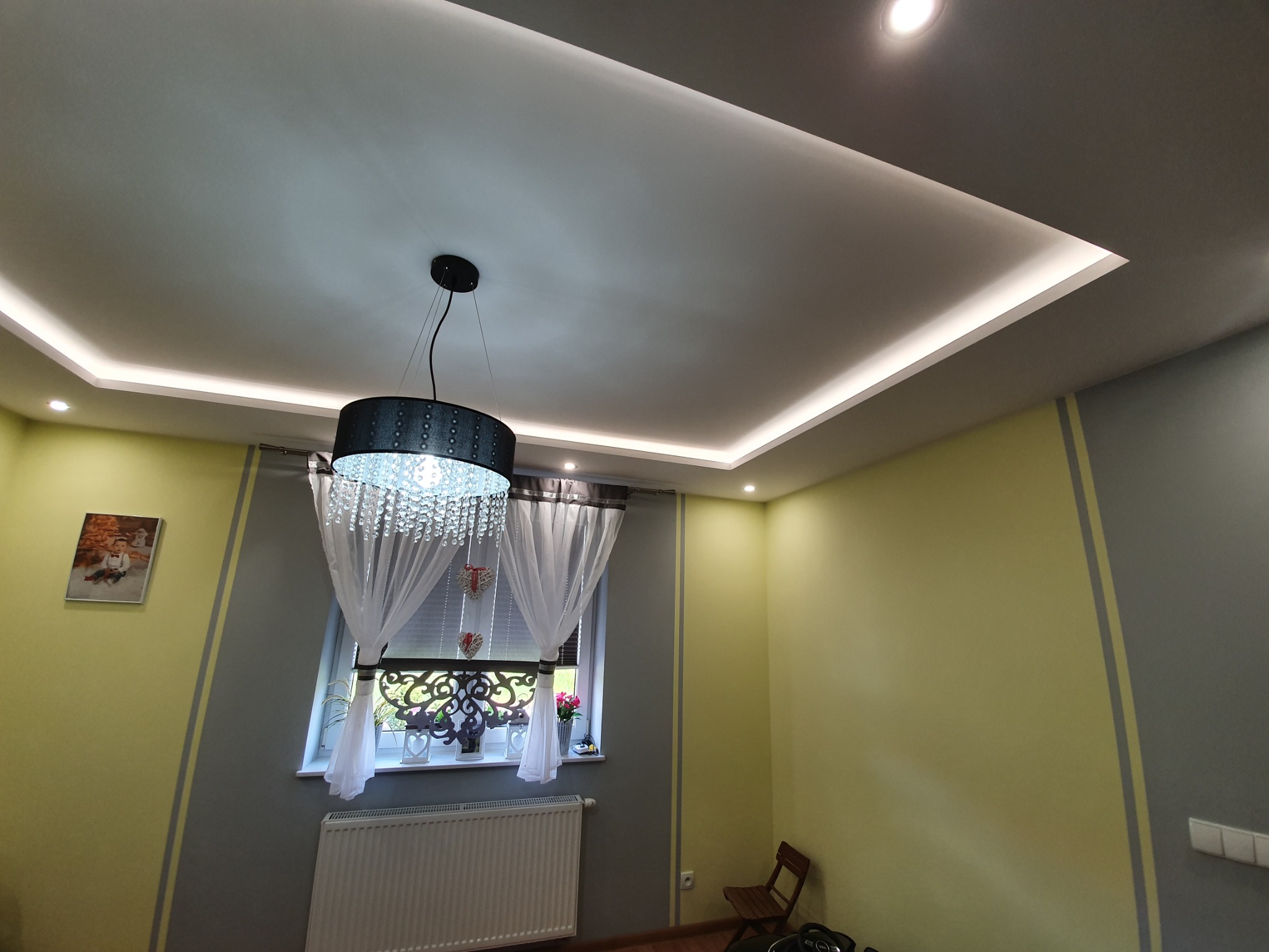 Wykończony pokój w Słopnicach z podwieszanym sufitem z oświetleniem LED, ozdobnym żyrandolem i ścianami w odcieniach żółci i szarości.