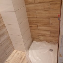 Sylmil - Łazienka w trakcie wykańczania: ułożone płytki imitujące drewno na ścianach i podłodze, obudowany pion rur płytkami w odcieniu beżu, brodzik zabezpieczony folią ochronną.