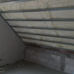 Poddasze z widoczną konstrukcją metalową i pianą izolacyjną, surowy stan wykończenia ścian i podłogi, przewody elektryczne na podłodze.