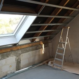 Sylmil - Poddasze w trakcie adaptacji: widoczne metalowe profile sufitowe, okno dachowe, fragment ściany z betonu komórkowego i drabina aluminiowa. Na podłodze leżą dodatkowe profile.