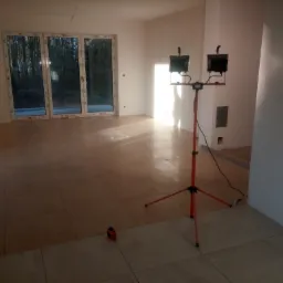 Wnętrze w trakcie układania płytek podłogowych, widoczne płytki w odcieniach beżu, lampa budowlana na trójnogu i dalmierz laserowy.