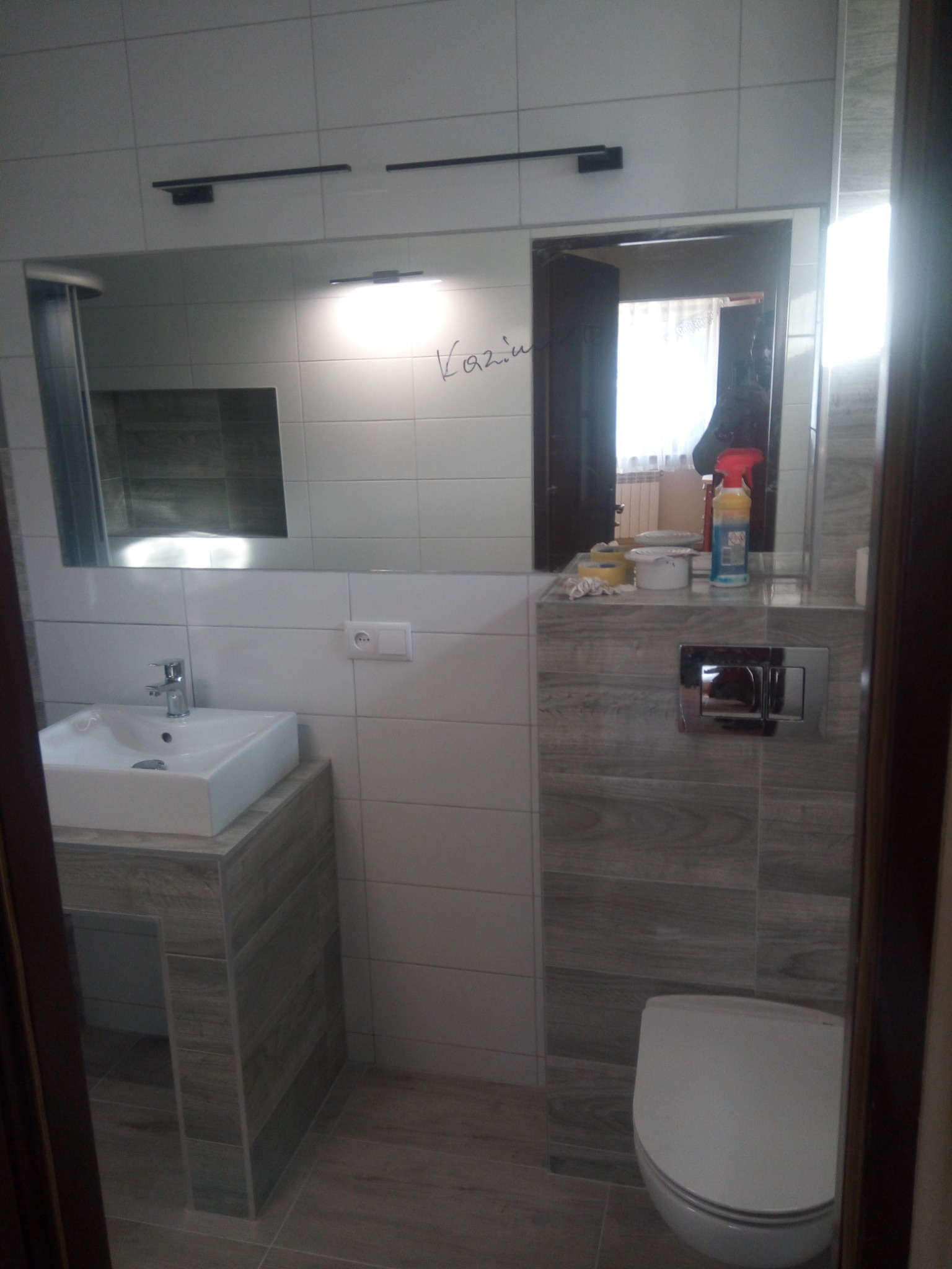 Wykończona łazienka z białymi płytkami, umywalką na postumencie z płytek imitujących drewno oraz toaletą. W lustrze odbija się część pokoju z oknem.