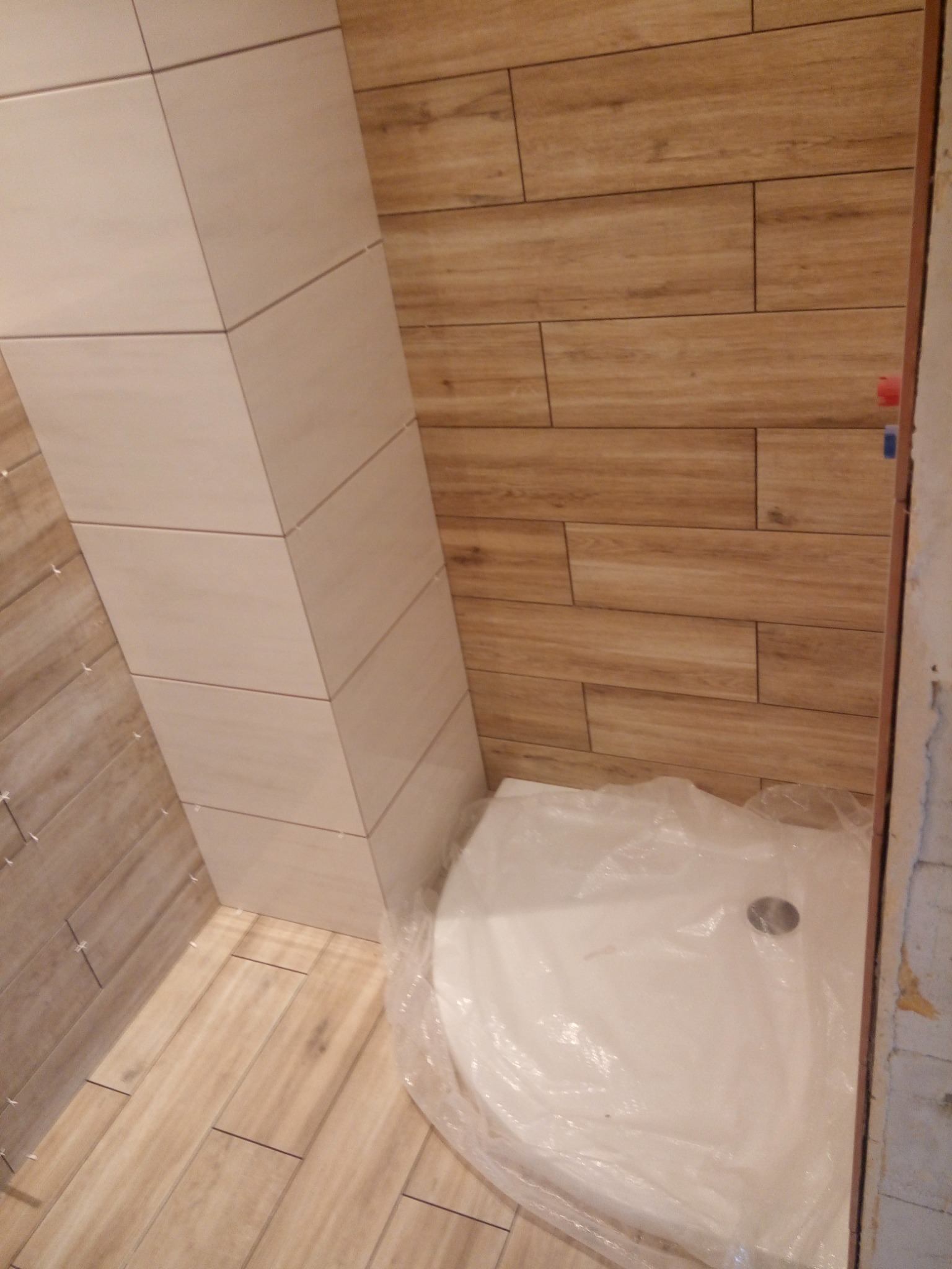 Łazienka w trakcie wykańczania: ułożone płytki imitujące drewno na ścianach i podłodze, obudowany pion rur płytkami w odcieniu beżu, brodzik zabezpieczony folią ochronną.