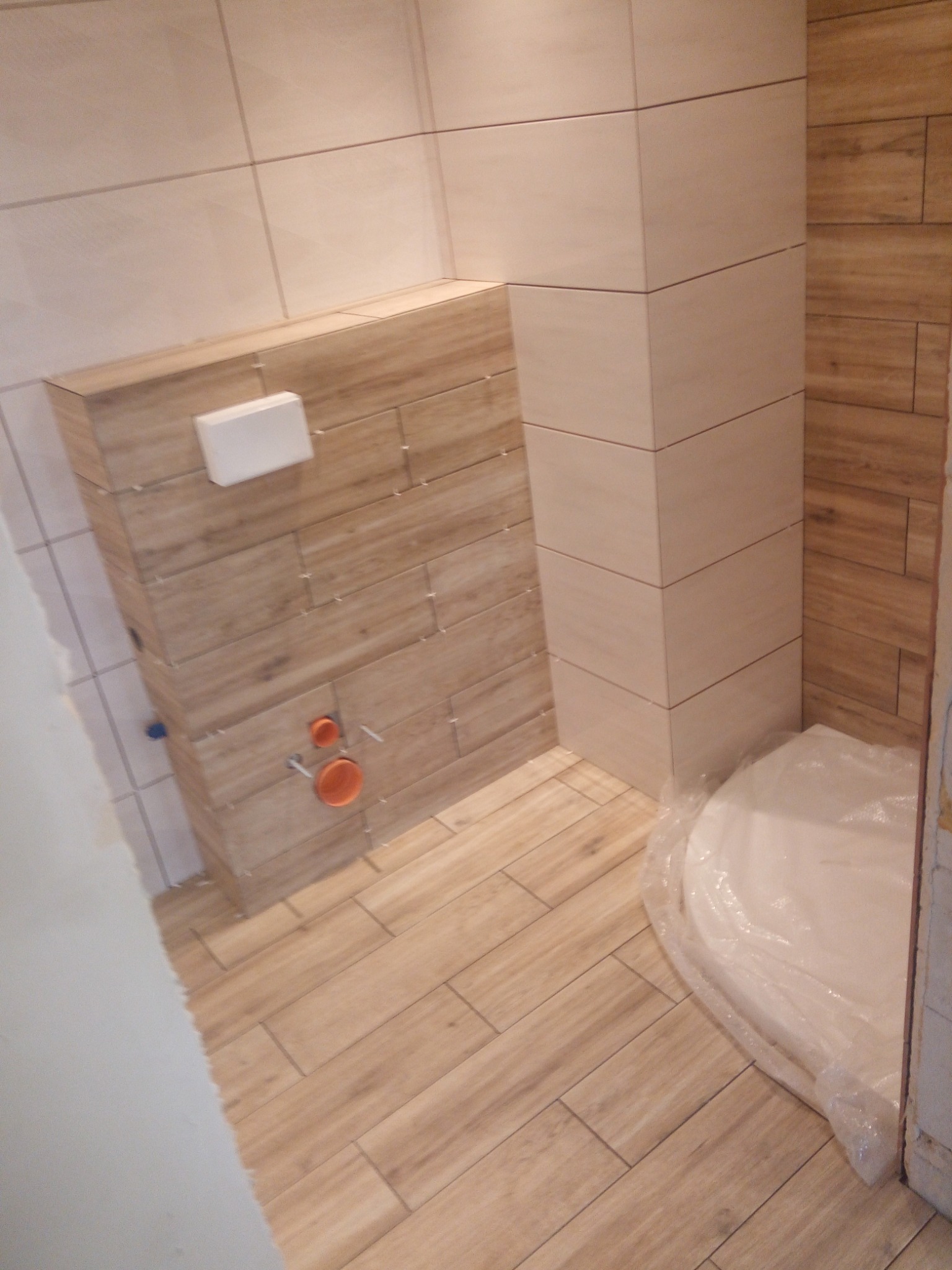 Nowoczesna łazienka w trakcie wykańczania, z drewnopodobnymi płytkami na ścianach i podłodze, zabudową WC i zabezpieczonym folią brodzikiem.