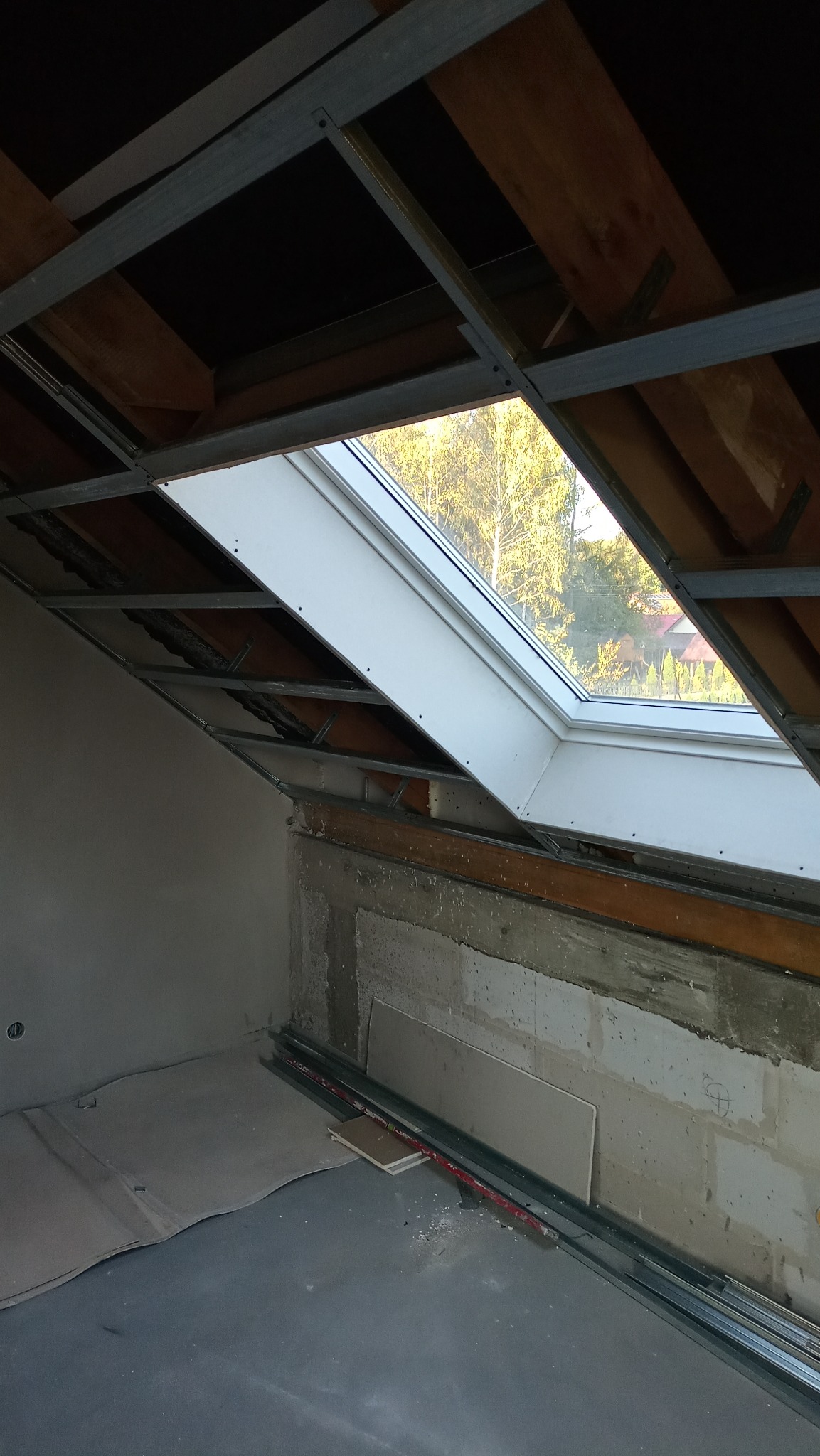 Poddasze w trakcie adaptacji: widoczne metalowe profile konstrukcyjne sufitu podwieszanego, okno dachowe z widokiem na drzewa, surowe ściany i podłoga przygotowane do dalszych prac wykończeniowych.