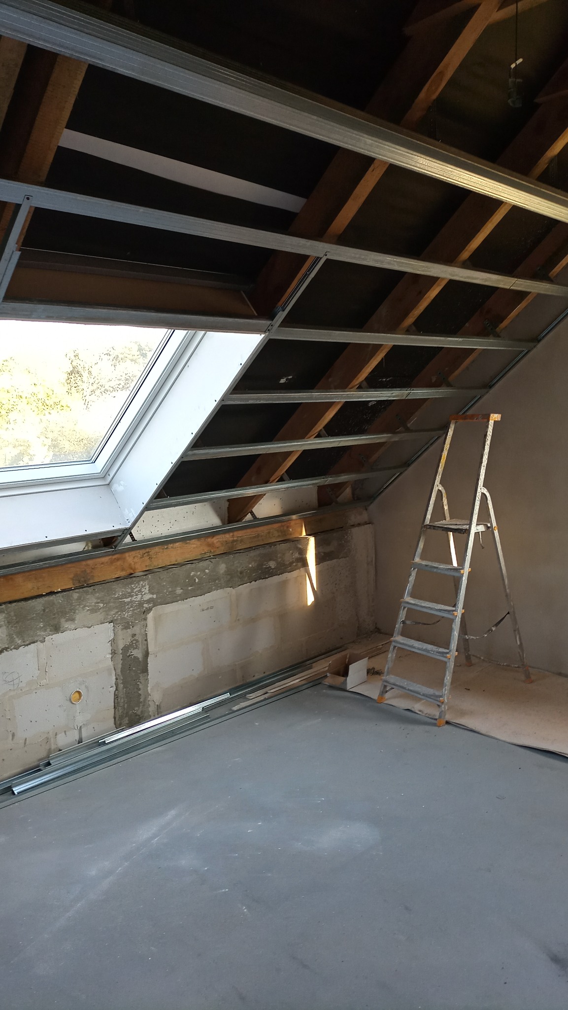 Poddasze w trakcie adaptacji: widoczne metalowe profile sufitowe, okno dachowe, fragment ściany z betonu komórkowego i drabina aluminiowa. Na podłodze leżą dodatkowe profile.