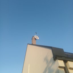 Instalacje antenowe sat TV DVB-T T2 - Antena satelitarna zamontowana na kominie domu jednorodzinnego, widok z dołu na tle błękitnego nieba.