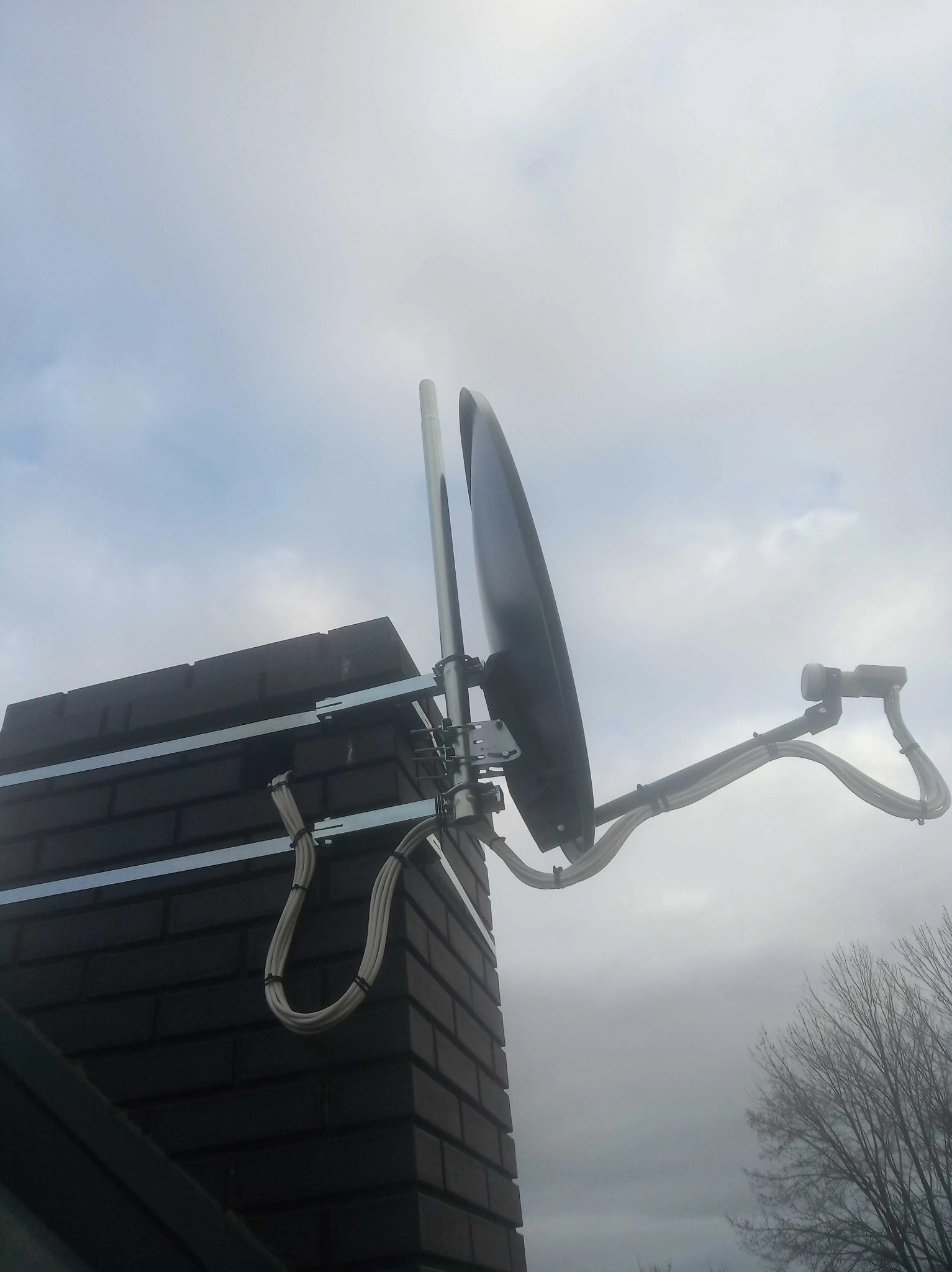 Antena satelitarna zamontowana na kominie z cegły, widok z dołu na tle pochmurnego nieba.