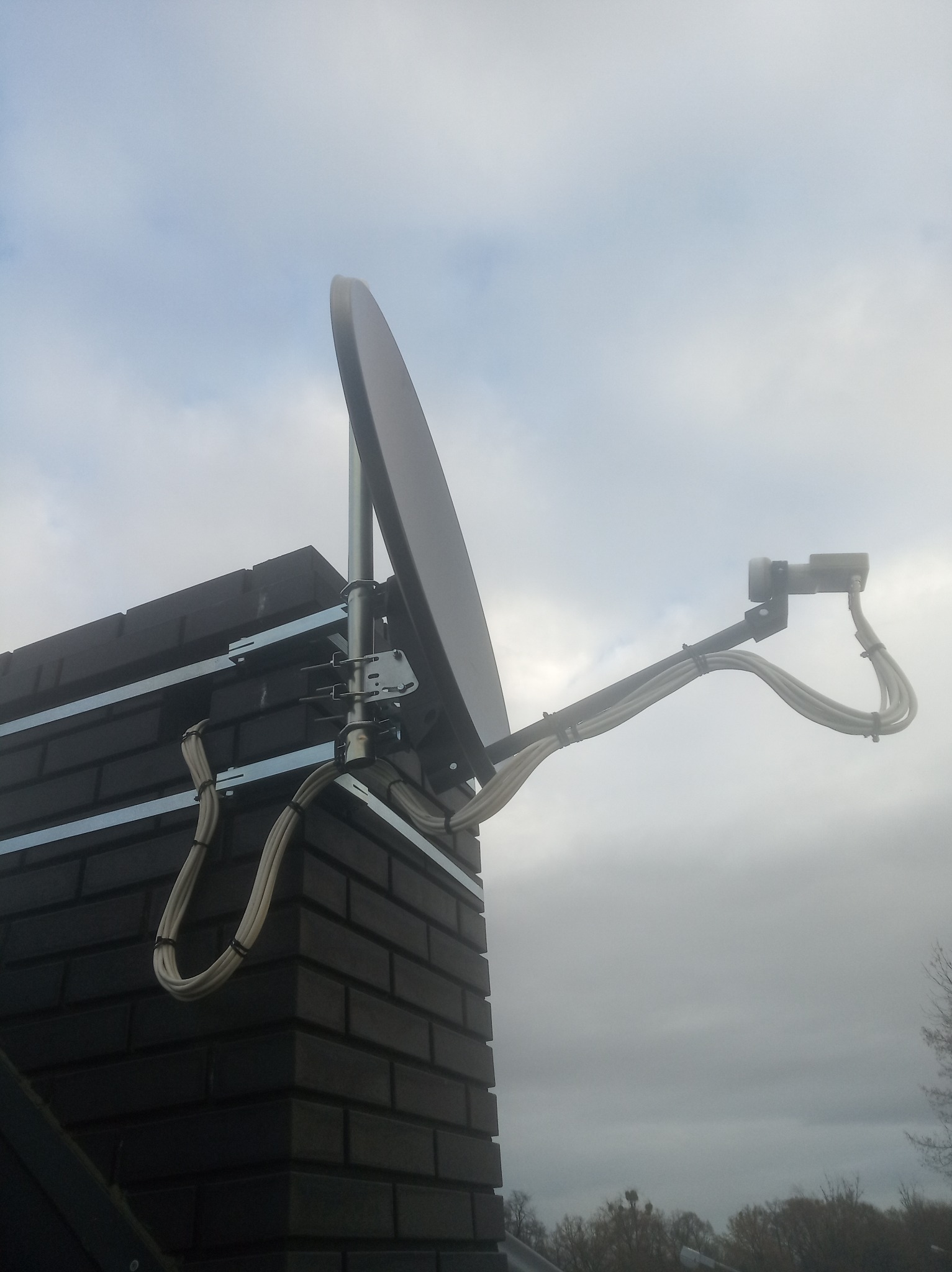 Antena satelitarna zamontowana na kominie z ciemnej cegły, kable starannie poprowadzone i przymocowane, pochmurne niebo w tle.
