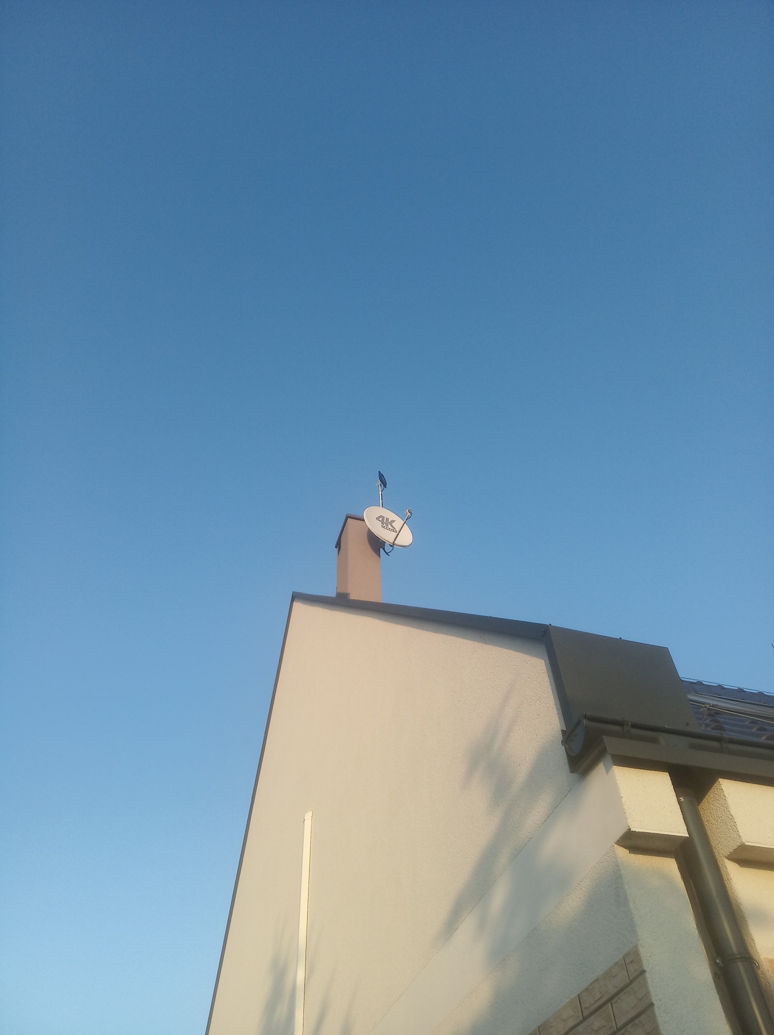 Antena satelitarna zamontowana na kominie domu jednorodzinnego, widok z dołu na tle błękitnego nieba.