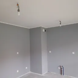 Świeżo pomalowane na szaro wnętrze pokoju w Legnicy z widocznymi gniazdkami elektrycznymi i wyprowadzonymi rurami instalacji wodnej, oświetlone tymczasowymi żarówkami.