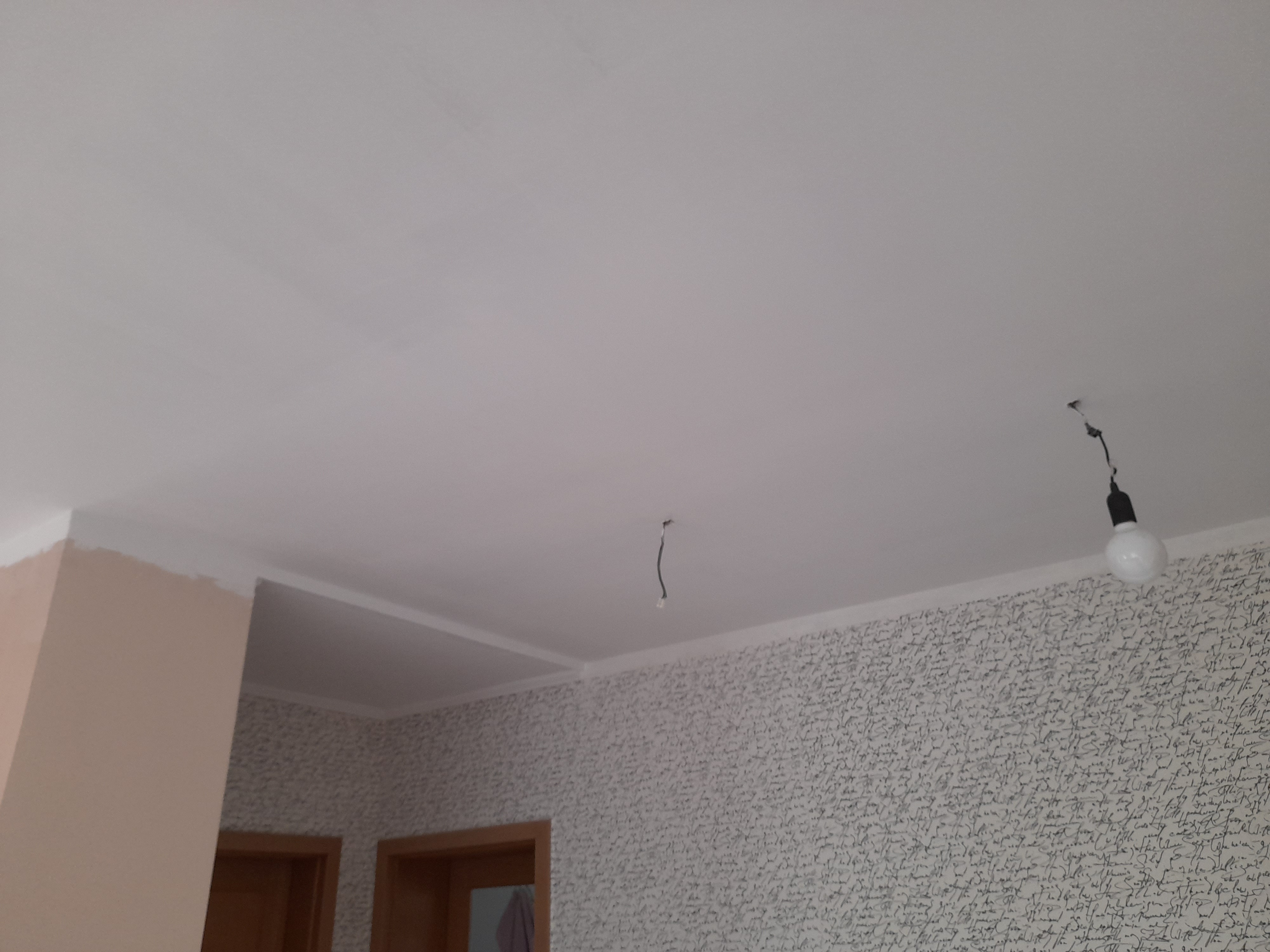 Pomalowany na biało sufit w mieszkaniu z widocznymi przewodami od oświetlenia i fragmentem ściany z tapetą w stylu retro oraz pomalowanym fragmentem ściany przy drzwiach.