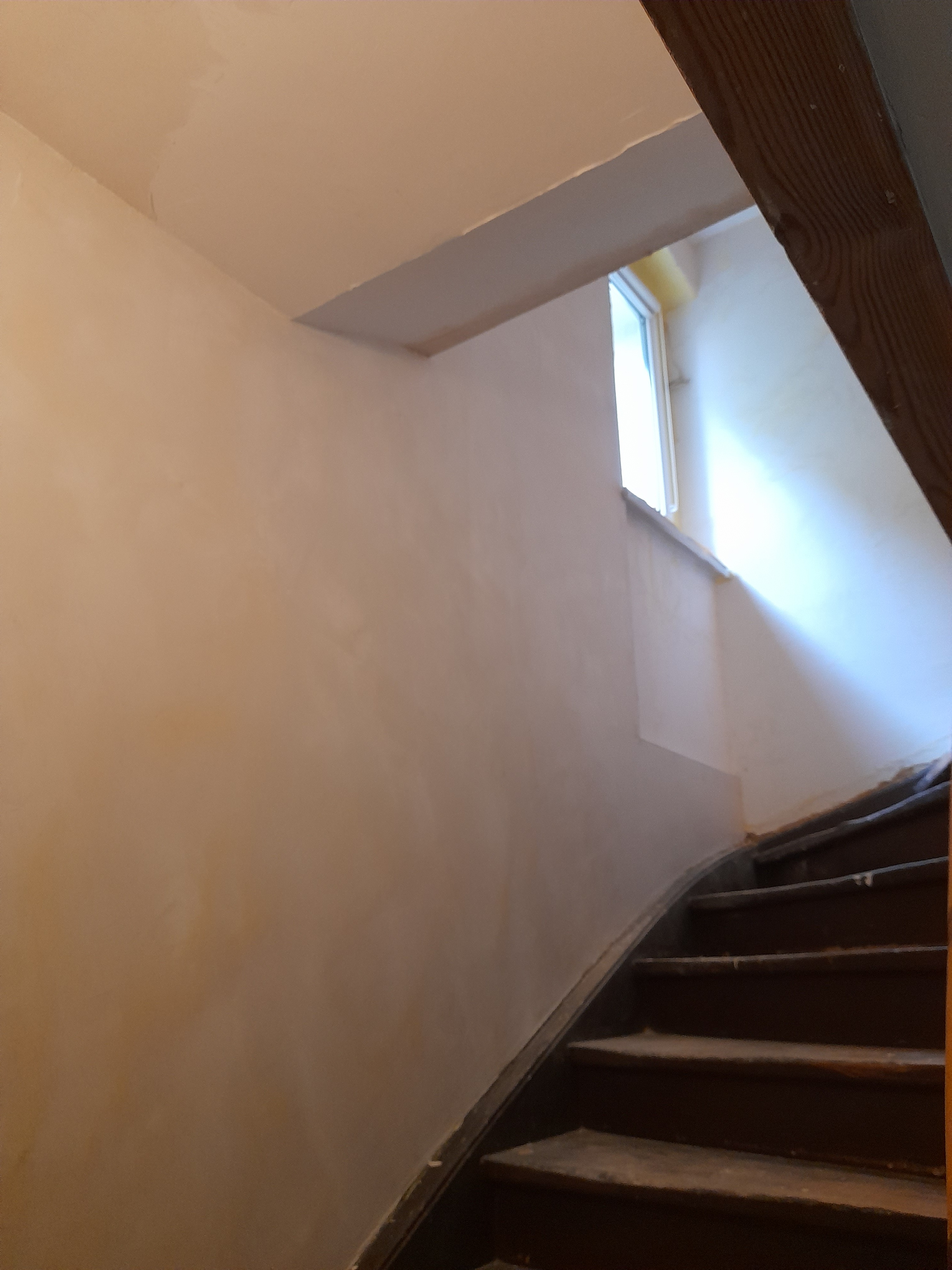 Ściana po szpachlowaniu na klatce schodowej, widoczne ślady po gładzi, okno z białymi ramami w tle.