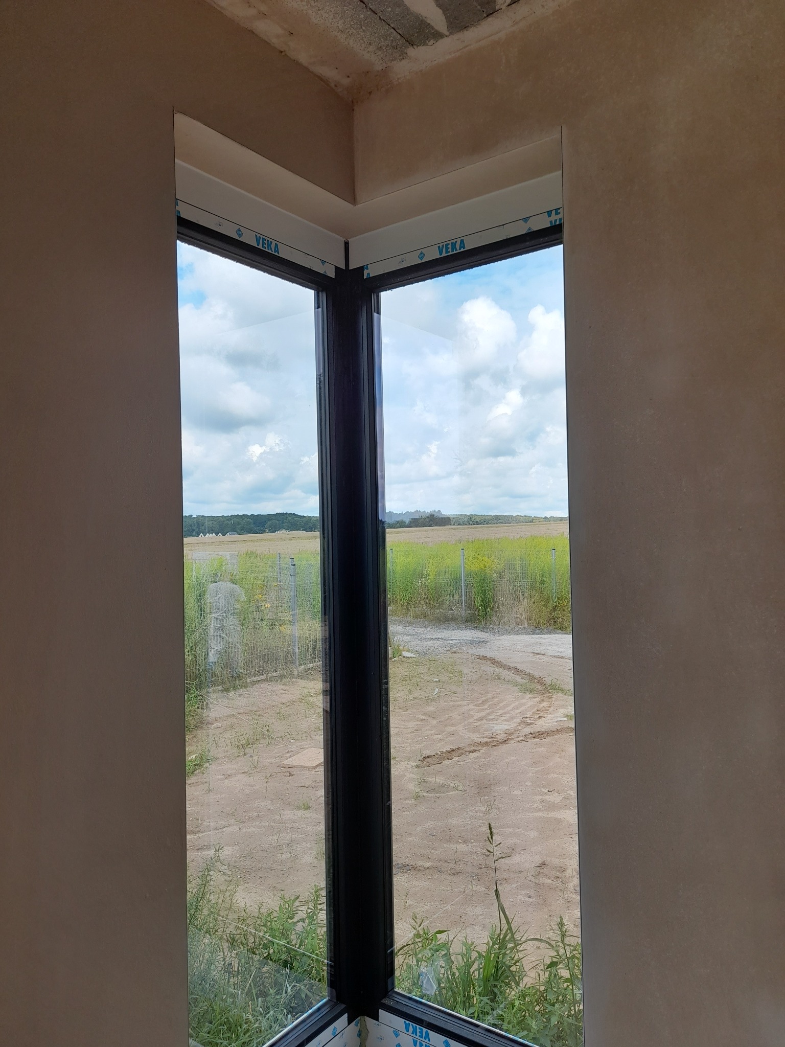 Fragment wnętrza z narożnym oknem, widok na pole i niebo, ściany w trakcie wykończenia, widoczne ślady po szpachlowaniu.