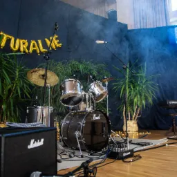 Scena z perkusją Pearl, keyboardem Yamaha, wzmacniaczem Marshall i dekoracją z roślin, oświetlona światłami i ozdobiona napisem z balonów 'COOLTURALYE'.