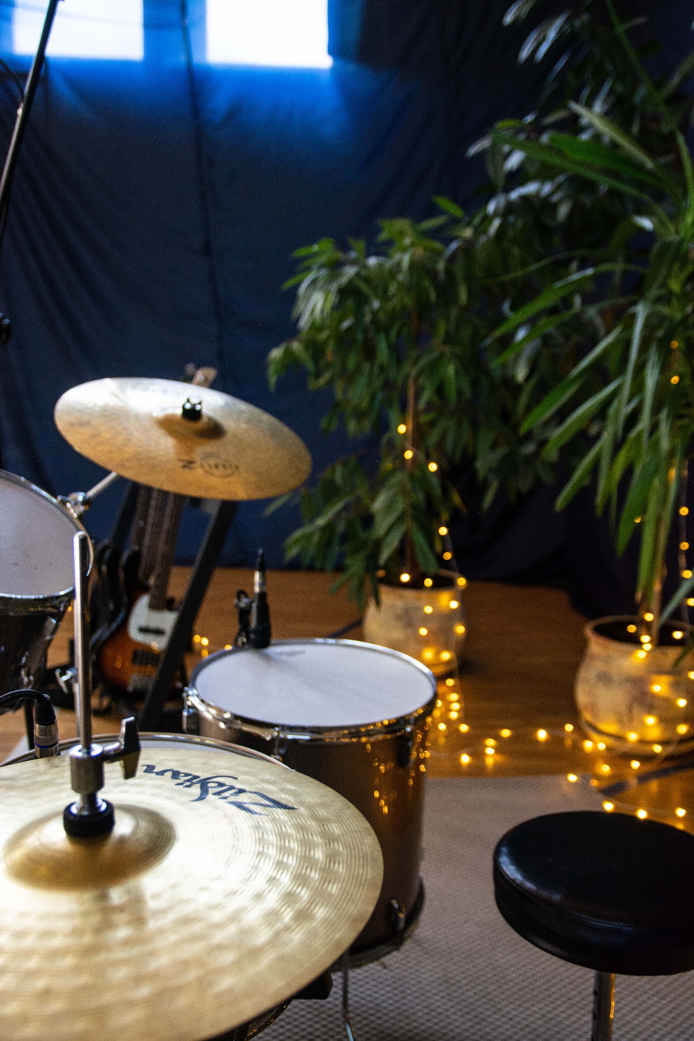 Fragment zestawu perkusyjnego z talerzami Zildjian, basówką w tle i roślinami ozdobionymi lampkami, w pomieszczeniu z ciemnym tłem.
