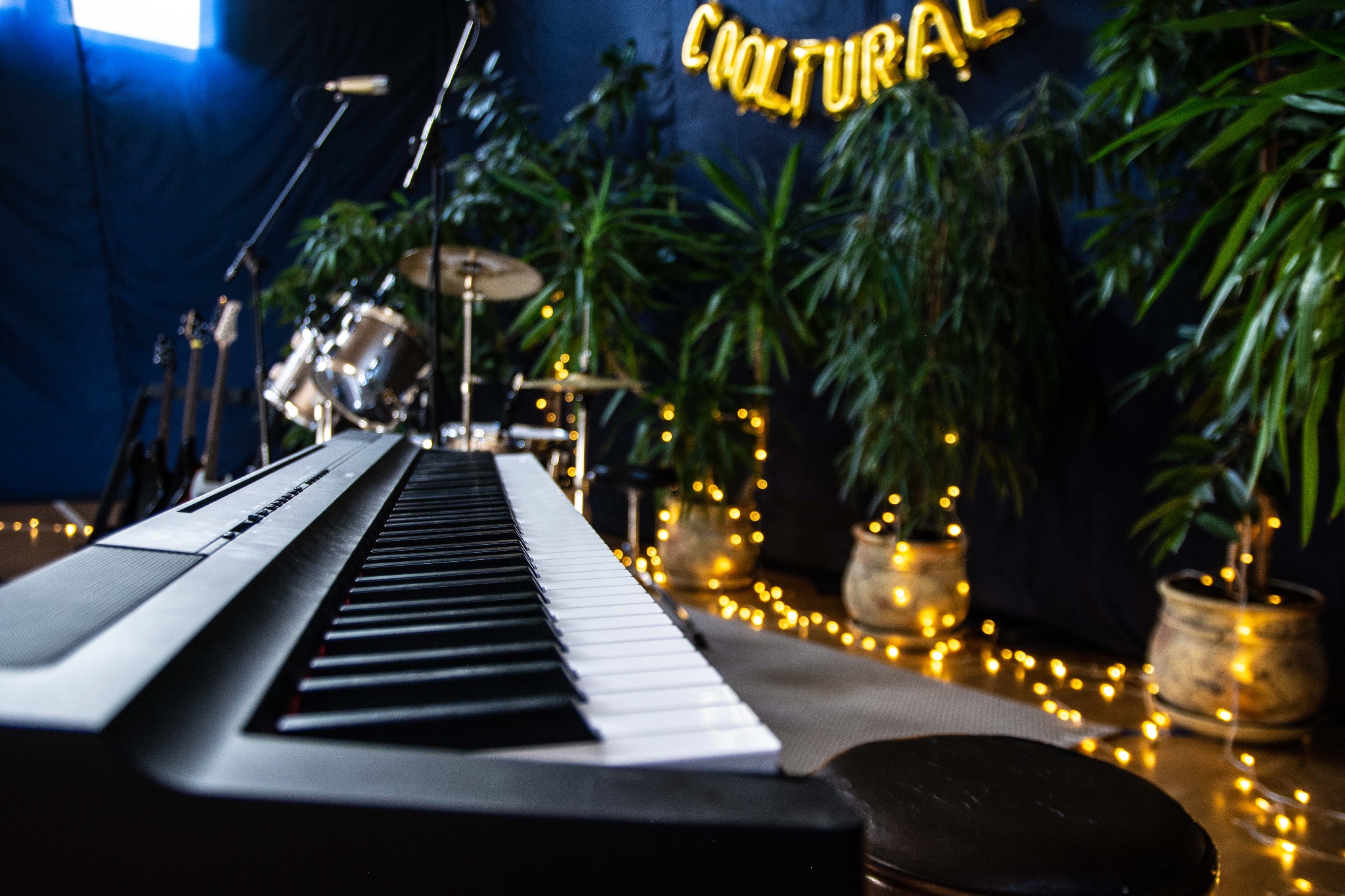 Klawiatura pianina cyfrowego na pierwszym planie, w tle perkusja, gitary, rośliny w doniczkach i napis 'COOLTURAL' z balonów, całość oświetlona dekoracyjnymi lampkami.