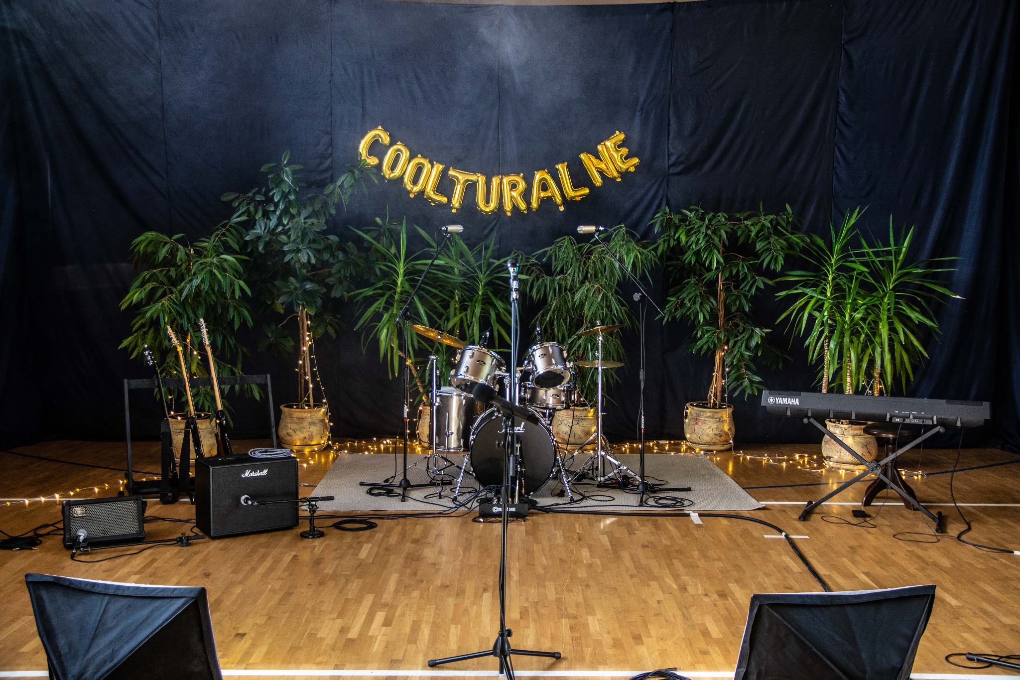 Scena muzyczna z perkusją, gitarami, keyboardem Yamaha, wzmacniaczami Marshall i Roland, mikrofonami, roślinami w donicach i złotymi balonami z napisem 'COOLTURALNE' na czarnym tle.