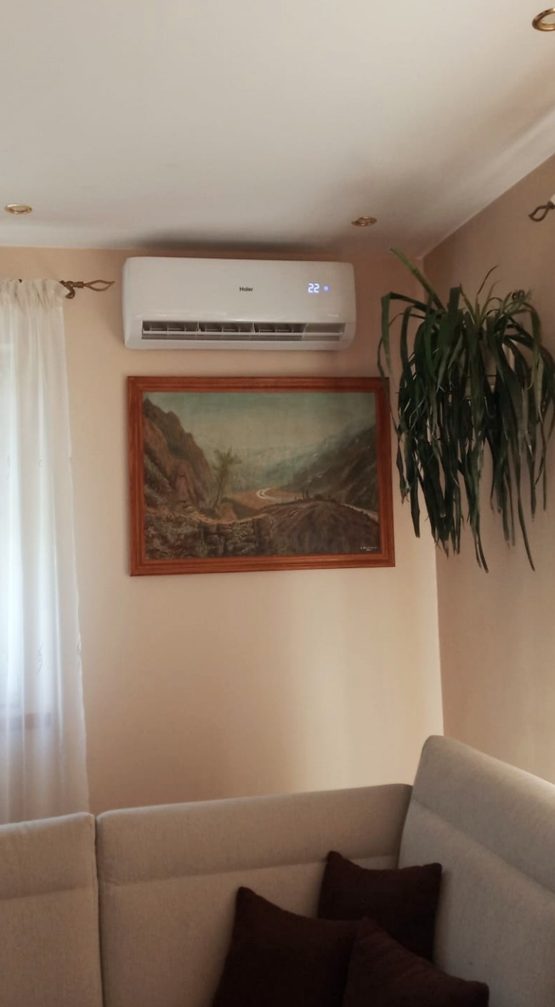 Biała klimatyzacja Haier z wyświetlaczem temperatury 22 stopnie, zamontowana nad obrazem z górskim krajobrazem w drewnianej ramie, obok zwisająca roślina doniczkowa, widok z narożnika kremowej sofy...
