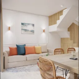 Jasne wnętrze salonu z jadalnią: beżowa sofa z kolorowymi poduszkami, drewniany stół z rattanowymi krzesłami, schody z drewnianymi panelami, obraz z motywem morskim.