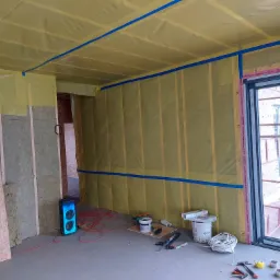 Wnętrze w trakcie prac: ściany i sufit pokryte wełną mineralną i folią paroizolacyjną, zabezpieczone niebieską taśmą malarską, widoczne narzędzia i materiały budowlane, głośnik przenośny na podłodze.