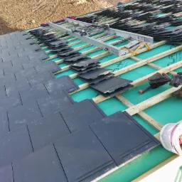 Montaż czarnej dachówki na dachu w Koszalinie, widoczna drabina dekarska, narzędzia i wiadro z materiałami uszczelniającymi VELUX.