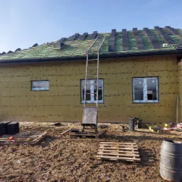 Dom jednorodzinny w trakcie prac dekarskich i ocieplania elewacji, widoczne ułożone dachówki, rusztowanie, ocieplenie ścian zewnętrznych wełną mineralną oraz przygotowane materiały budowlane na ziemi.