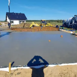Wylewka betonowa fundamentu domu jednorodzinnego podczas prac, widoczny betonowóz z pompą, koparka oraz pracownicy zacierający powierzchnię betonu.