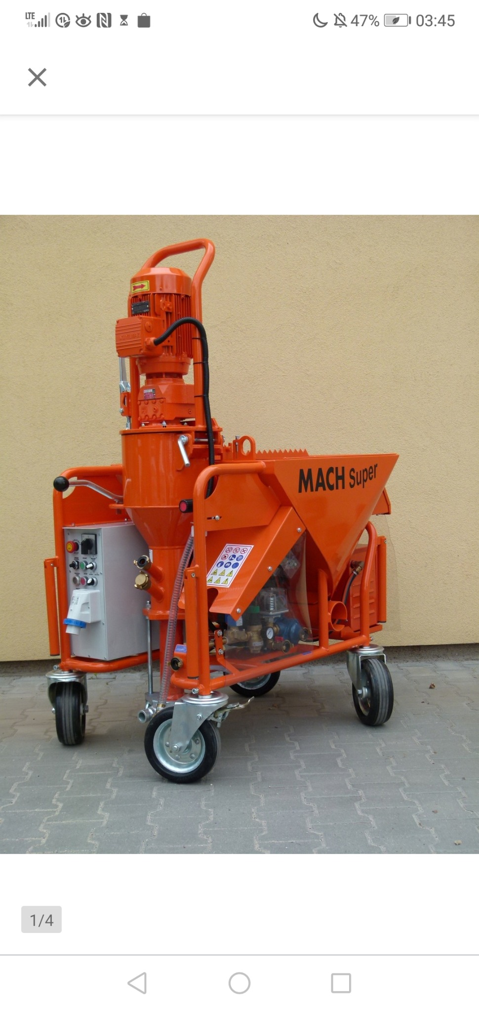 Pomarańczowa maszyna do tynków z napisem 'MACH Super' na kółkach, zaparkowana na kostce brukowej przed beżową ścianą.