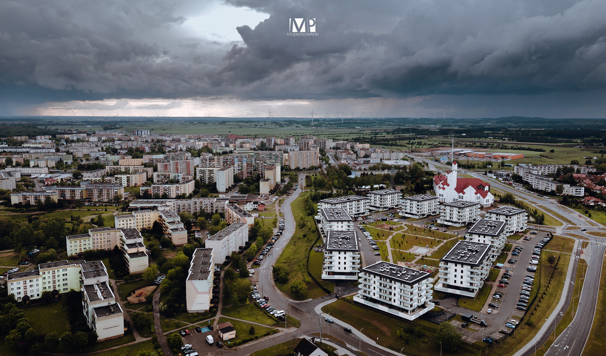 Panoramiczny widok z lotu ptaka na miasto z nowoczesnymi blokami mieszkalnymi, kościołem z czerwoną dachówką i turbinami wiatrowymi na horyzoncie pod burzowym niebem.