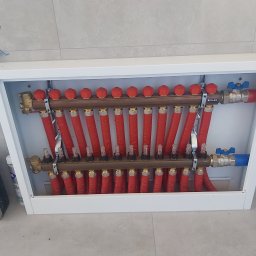 Maarrio Mariusz Piaskowski - Rozdzielacz podłogowy z czerwonymi rurami w białej szafce, widoczne zawory i liczniki przepływu, zamontowany na tle jasnych płytek.