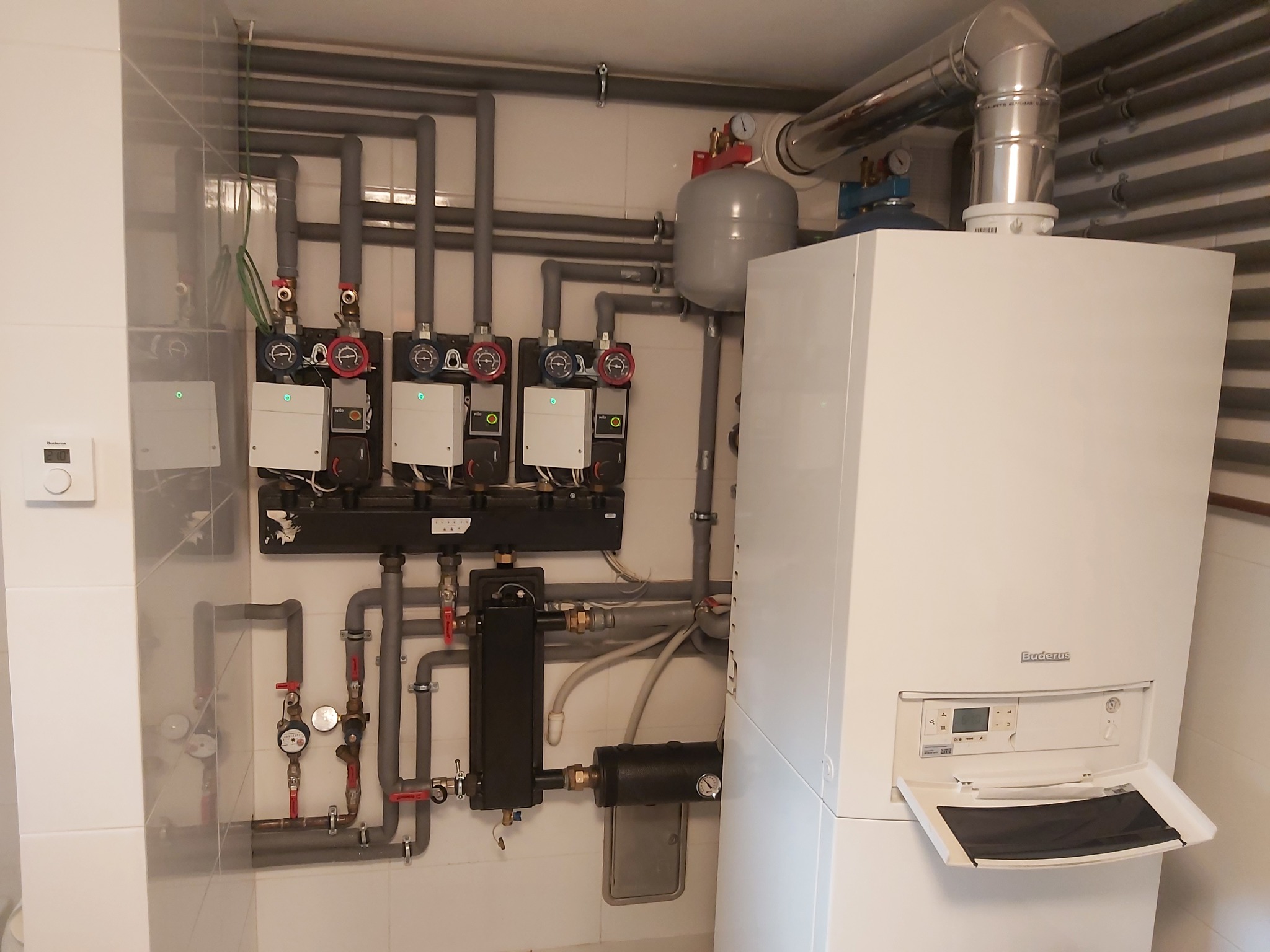 Biały piec grzewczy marki Buderus z otwartą klapką, podłączony do instalacji z szarymi rurami, czarnymi filtrami i rozdzielaczem z pompami obiegowymi w pomieszczeniu z białymi płytkami.