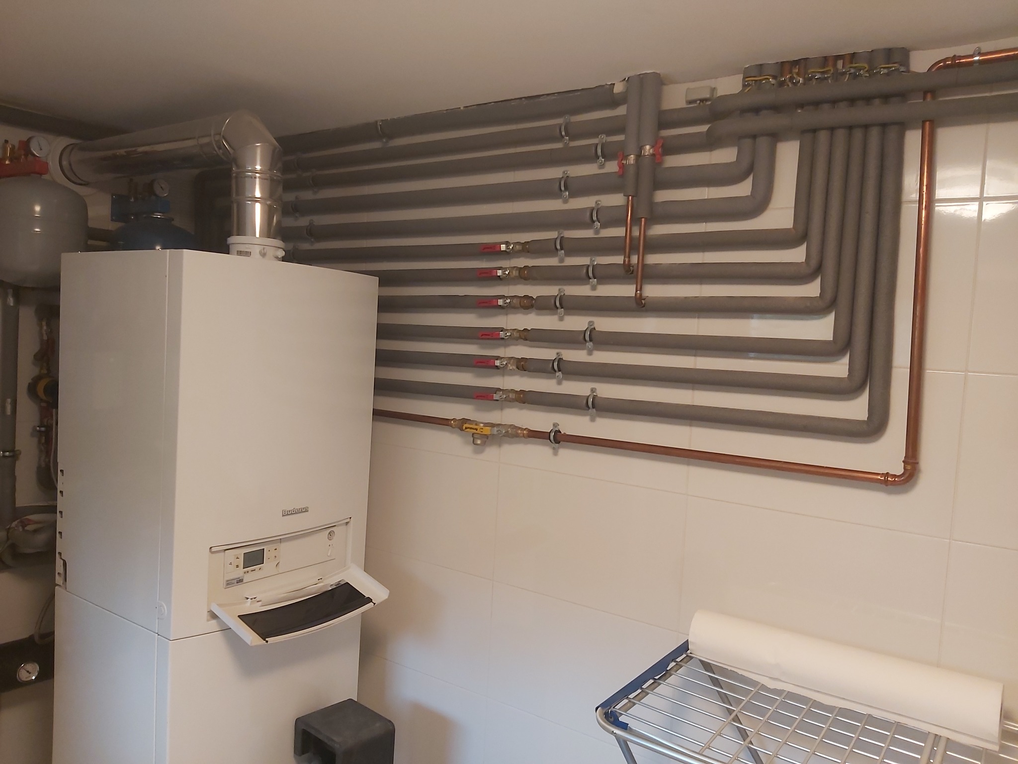 Biały piec grzewczy Buderus z otwartą klapką, widoczne liczne rury grzewcze zaizolowane szarą pianką, białe płytki na ścianie, fragment suszarki na pranie w rogu pomieszczenia.