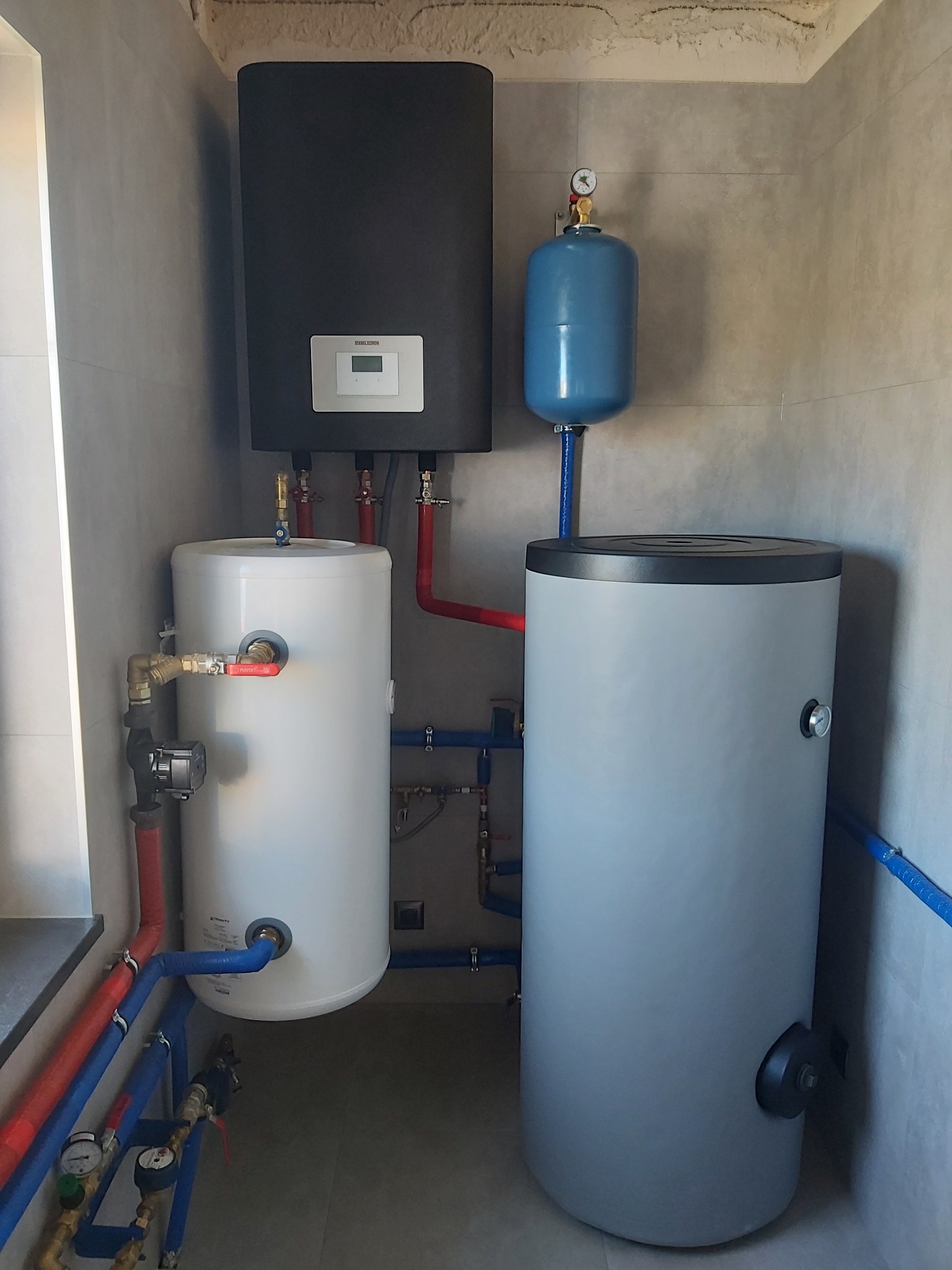 Kompaktowa kotłownia w rogu pomieszczenia, z czarnym kotłem wiszącym nad dwoma zbiornikami: białym i szarym, połączonymi rurami w kolorach czerwonym i niebieskim, z widocznymi manometrami i zaworami.
