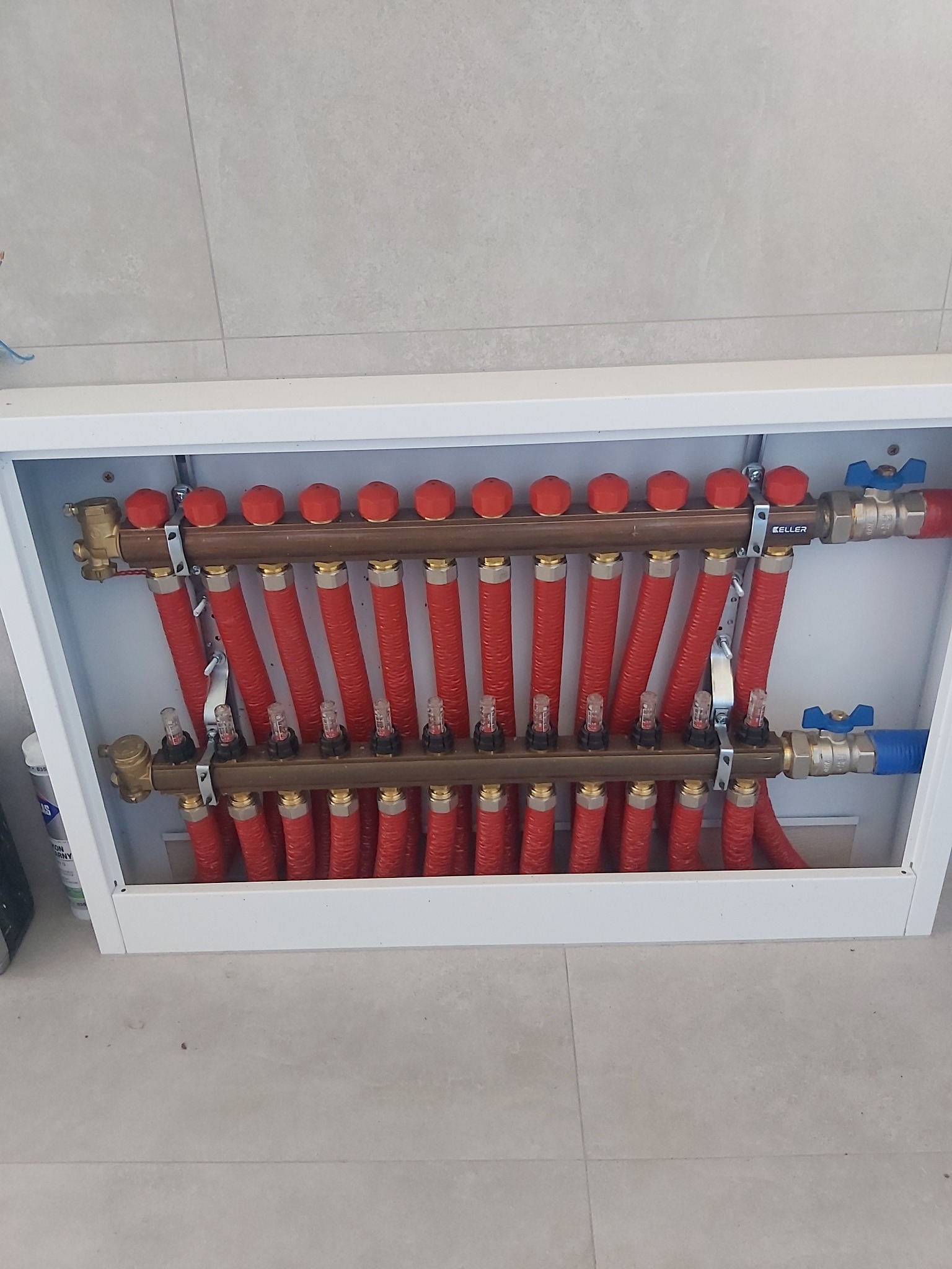 Rozdzielacz podłogowy z czerwonymi rurami w białej szafce, widoczne zawory i liczniki przepływu, zamontowany na tle jasnych płytek.