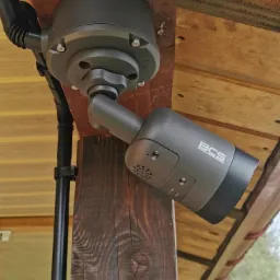 Szara kamera monitoringu BCS zamontowana na drewnianym słupie z widocznym okablowaniem w elastycznej rurze.