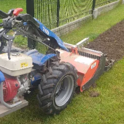 Glebogryzarka separacyjna Muratori, widok z boku, przygotowuje pas ziemi pod trawnik w ogrodzie z tujami i ogrodzeniem z siatki.