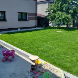 Świeżo założony trawnik z rolki w przydomowym ogrodzie, widoczny piaskownica z zabawkami i wózek dla lalek, drzewo w tle.