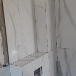 Ściana wyłożona białymi, marmurkowymi płytkami ceramicznymi, widoczna wnęka z otworem na element instalacyjny, fugi w kolorze szarym, w tle poziomica.