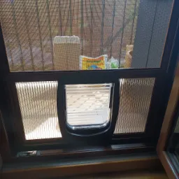 Brązowa rama z moskitierą i klapką dla zwierząt, widok na balkon z rattanowym krzesłem i podłogą z desek.