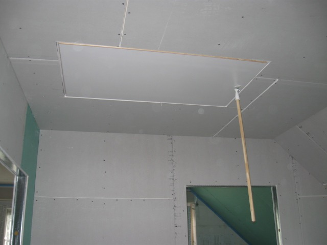 Montaż płyty gipsowo-kartonowej na poddaszu z widocznym otworem rewizyjnym i drewnianym drążkiem podtrzymującym klapę.