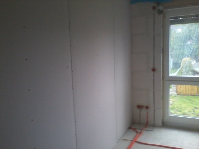 Wnętrze w trakcie wykańczania: ściany pokryte płytami gipsowo-kartonowymi, widoczne przewody elektryczne i okno z widokiem na deszczowy ogród.