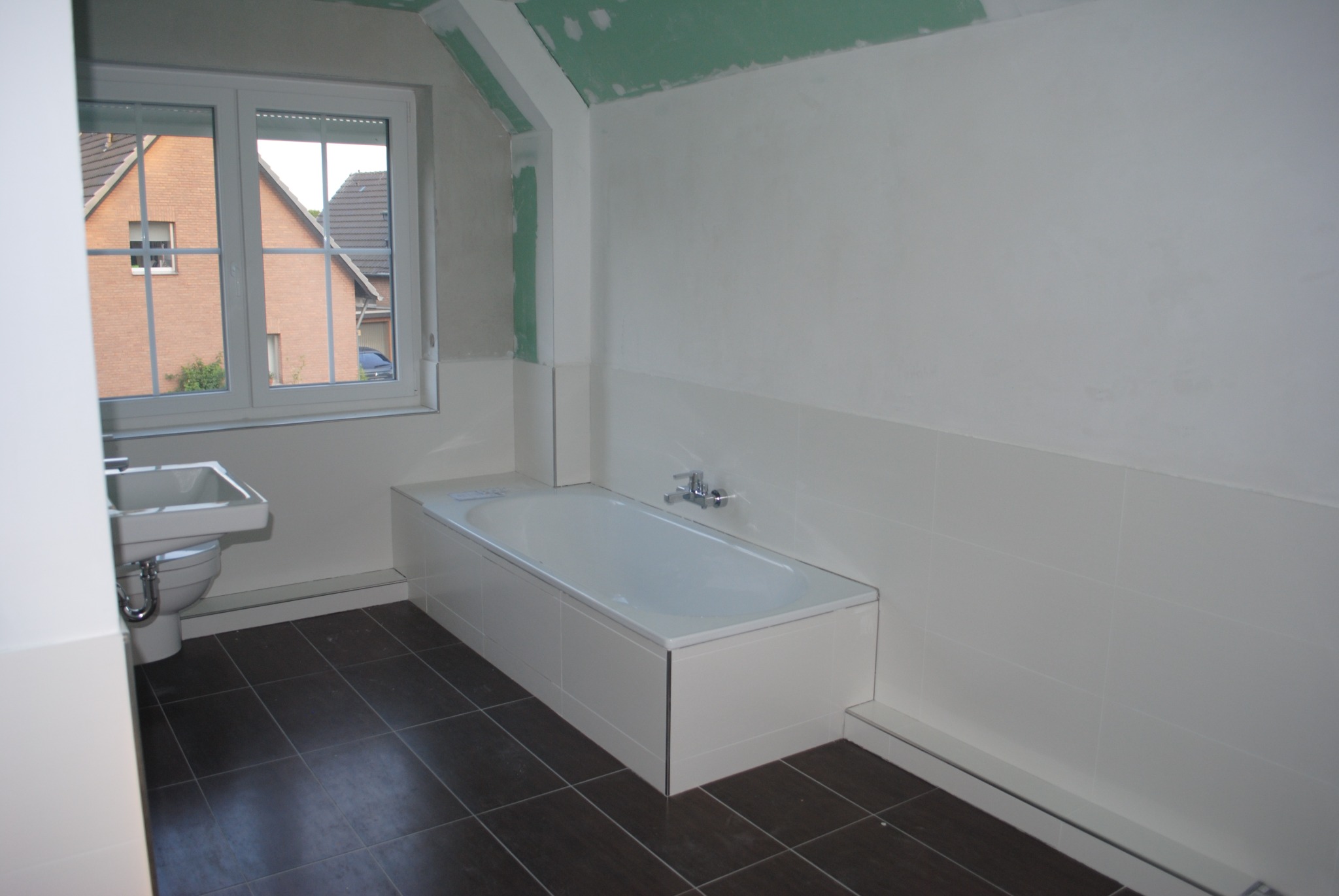 Łazienka w trakcie remontu: biała wanna obudowana płytkami, umywalka z toaletą, ciemne płytki podłogowe, ściany częściowo wykończone, sufit z widocznymi śladami szpachlowania, okno z widokiem...